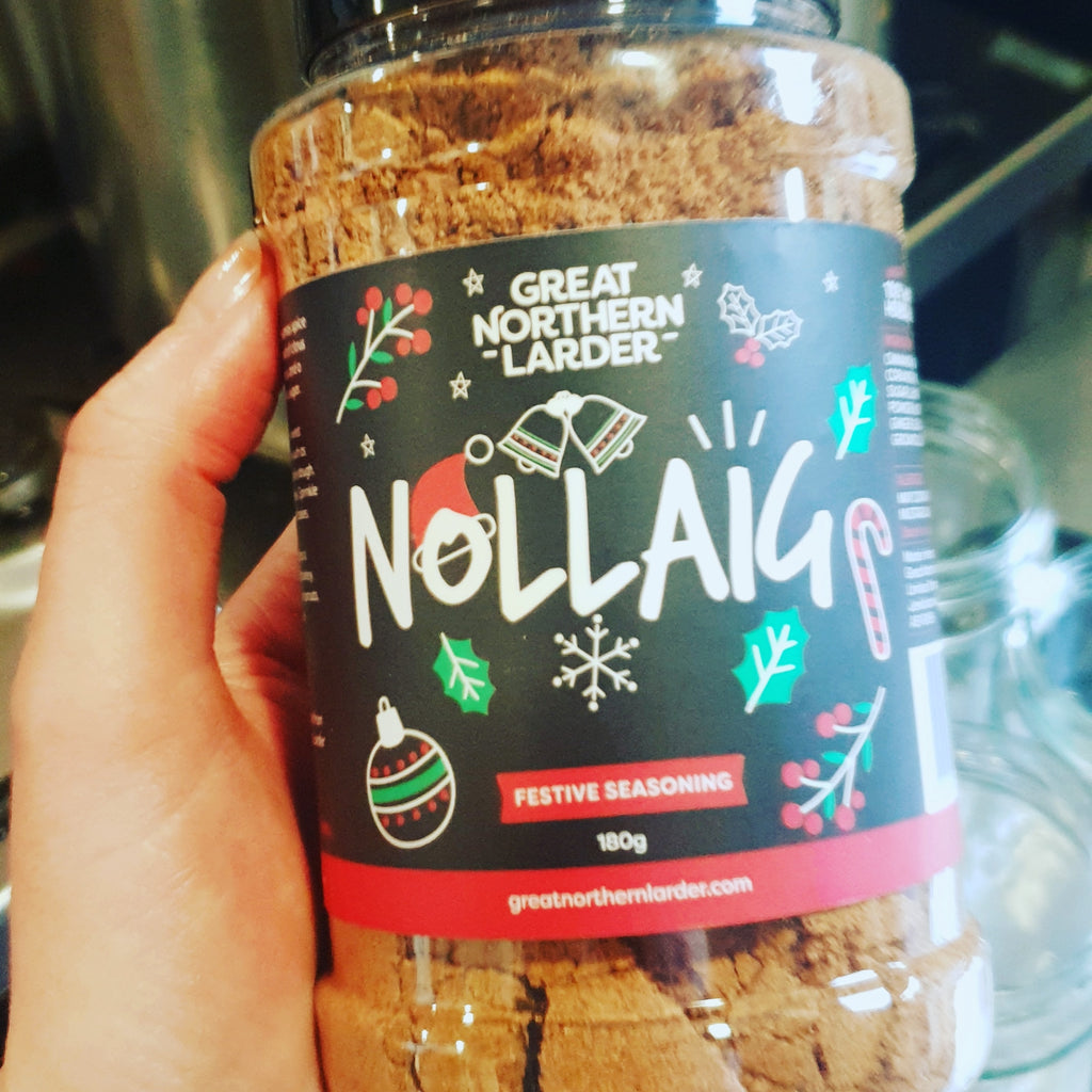A photograph of a tub of Nollaig spice being held in a hand. The label has outline of holly, berries, candy canes, baubles and snowflakes behind the name and logo.